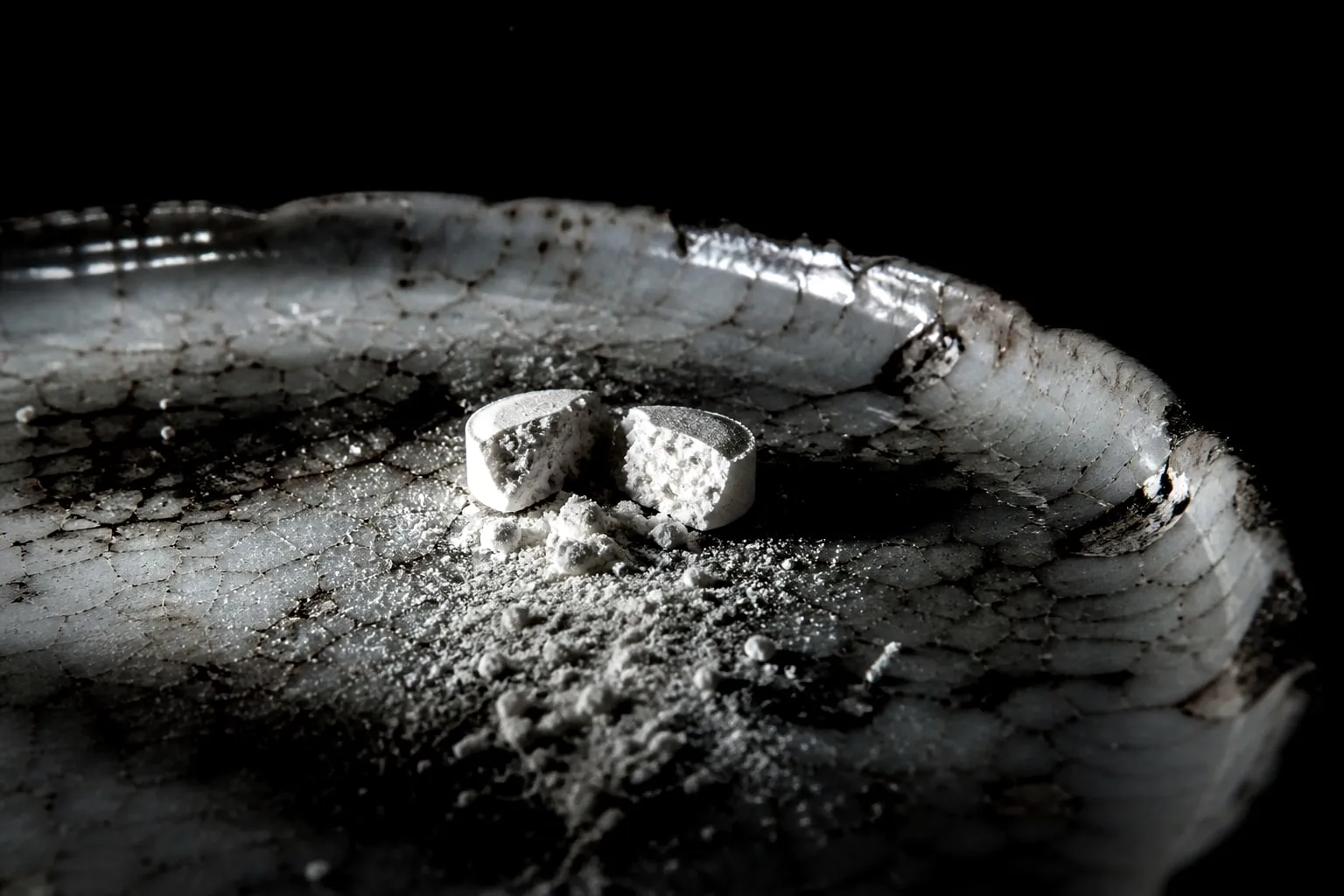 Editorial illustration: Split prescription tablet on a cracked porcelain saucer, its powder spilling like enamel dust un